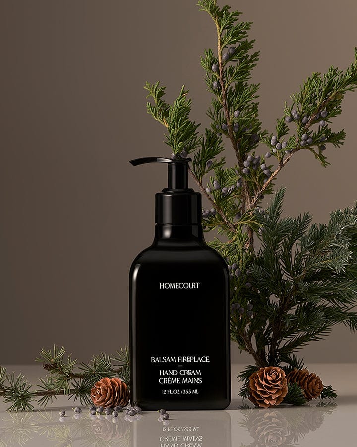 Black Homecourt Balsam Fireplace Hand Cream bottle with balsam branches and pine cones around it on a reflective surface.