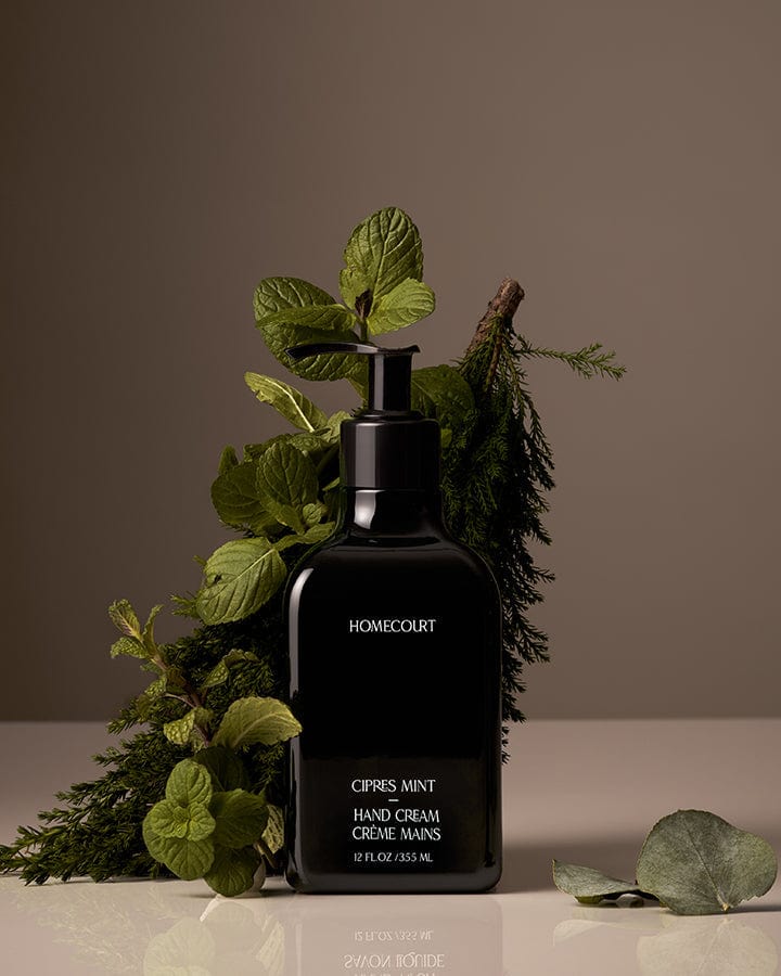 Black Homecourt Cipres Mint Hand Cream bottle with mint and cipres branches around it on a reflective surface.
