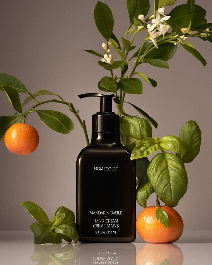 Black Homecourt Mandarin Hand Cream bottle with mandarin branches and oranges around it on a reflective surface.