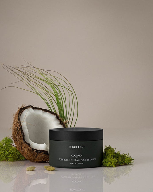 Black jar of Homecourt Indulgent Body Butter with a coconut and greenery on a neutral background.