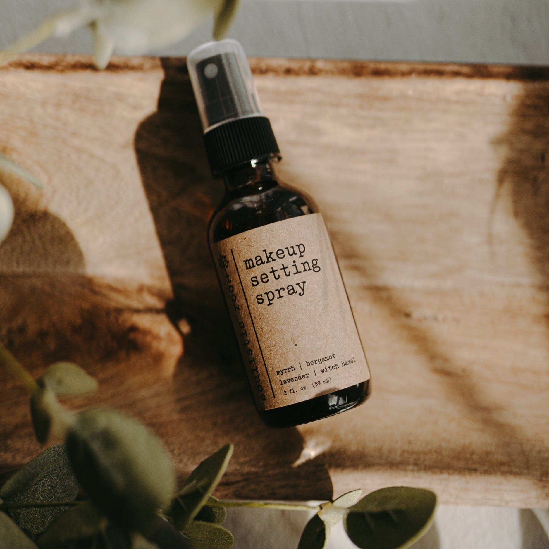 Makeup setting spray bottle with a label on a wooden surface with greenery.