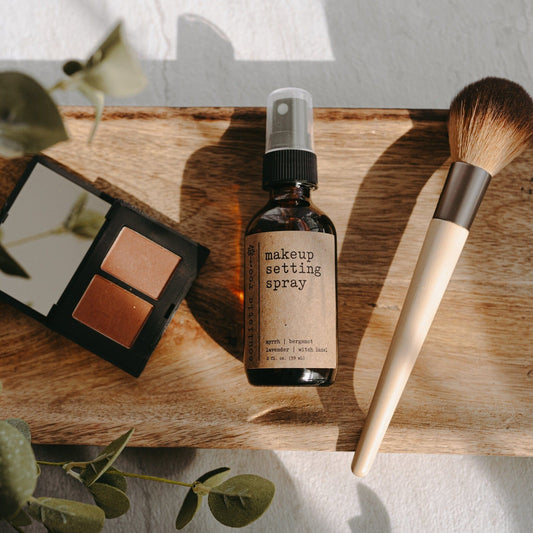 Makeup setting spray, brush, and color palette on a wooden board with greenery.