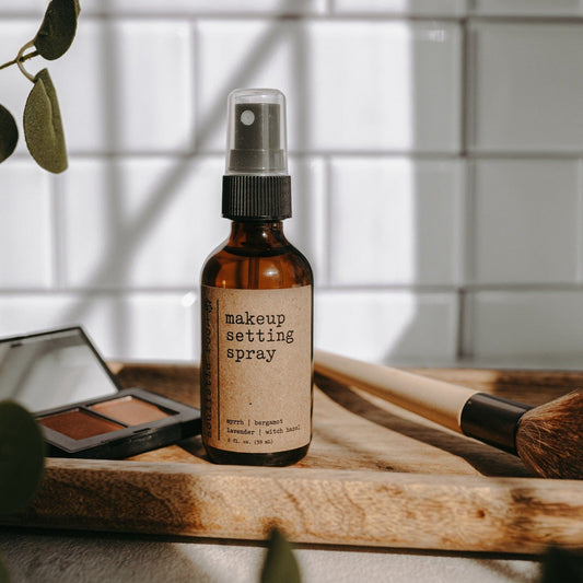 Makeup setting spray bottle on a wooden surface with blush in a bathroom with a plant and blurred background.