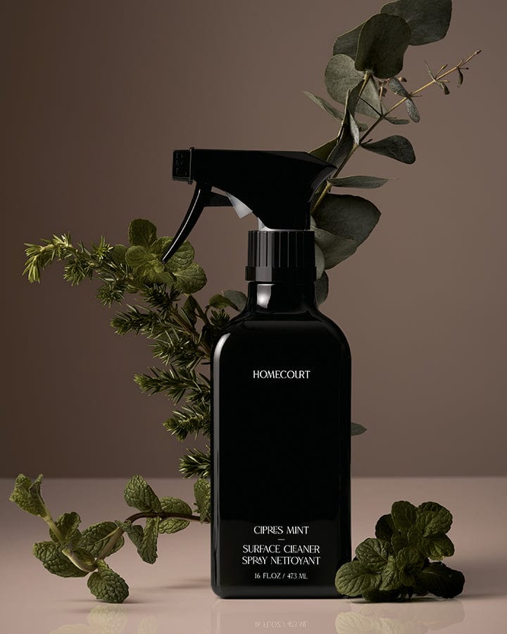 Black spray bottle of Homecourt Natural Surface Cleaner with green mint leaves on a brown background.