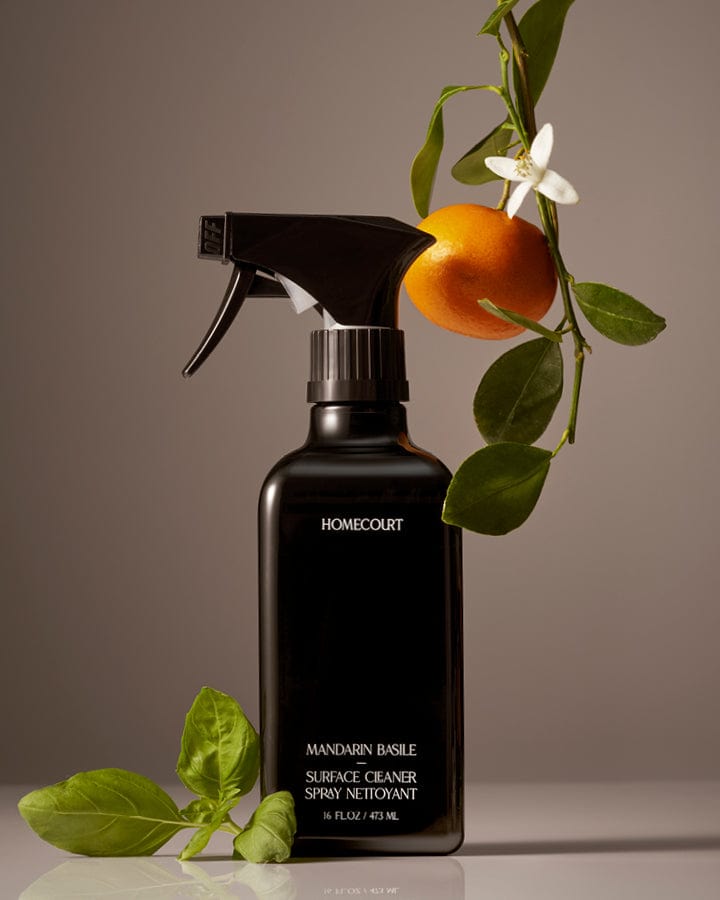 Black spray bottle of Homecourt Natural Surface Cleaner with mandarin orange and leaves on a neutral background.