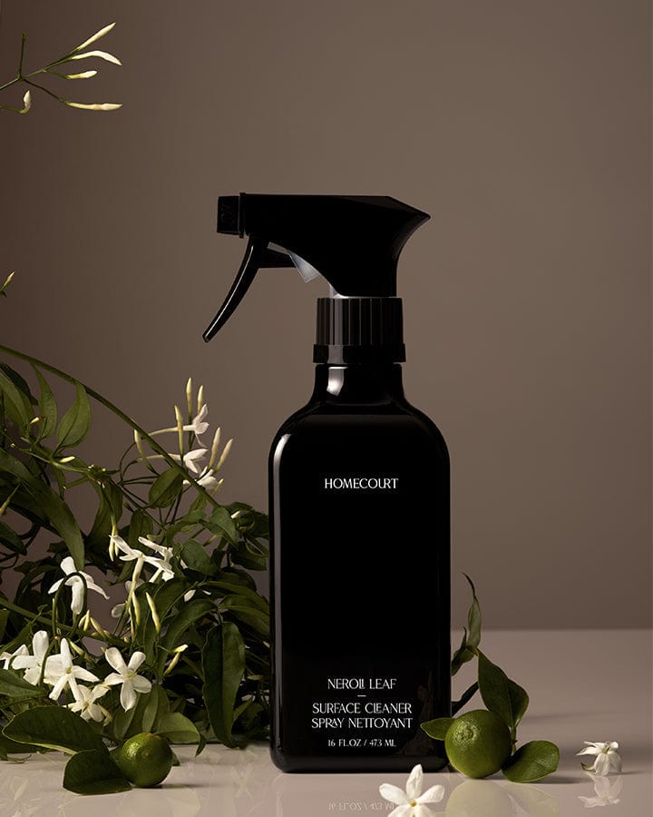 Black spray bottle of Homecourt Natural Surface Cleaner with green Neroli leaves and white flowers in the background.
