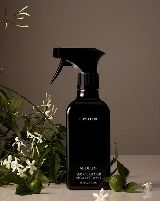 Black spray bottle of Homecourt Natural Surface Cleaner with green Neroli leaves and white flowers in the background.