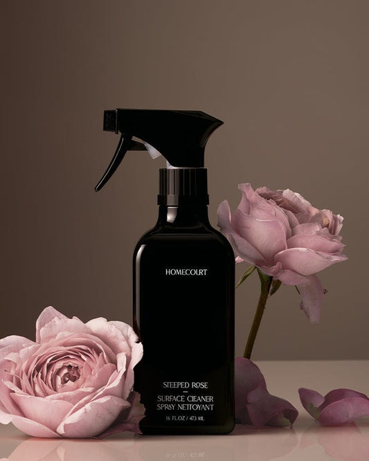 Black spray bottle of Homecourt Natural Surface Cleaner with pink roses on a brown background.