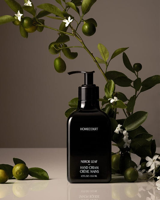 Black Homecourt Neroli Hand Cream bottle with neroli branches and fruit around it  on a reflective surface.