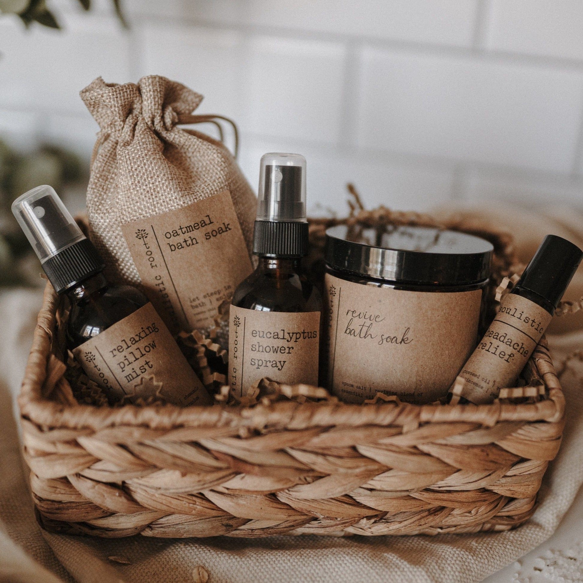 5-Piece Therapeutic Spa Gift Set of natural products in a basket on linen fabric with greenery in front of a tiled wall.
