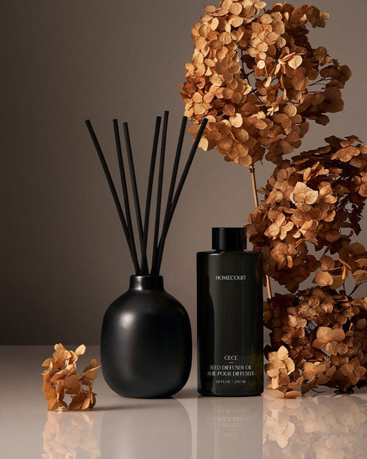 Black Reed diffuser with black sticks and a bottle of Home&Garden candle next to dried flowers on a reflective surface.